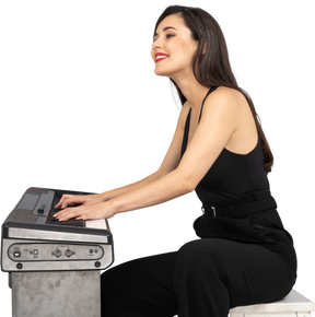 Side view of a smiling sitting young lady in black suit playing the piano