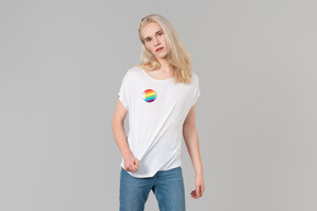 Good-looking young man with long blond hair, standing against grey background, wearing blue jeans and a white t-shirt with lgbt badge on it