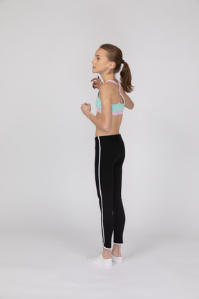 Three-quarter back view of a teen girl in sportswear raising hands and looking aside